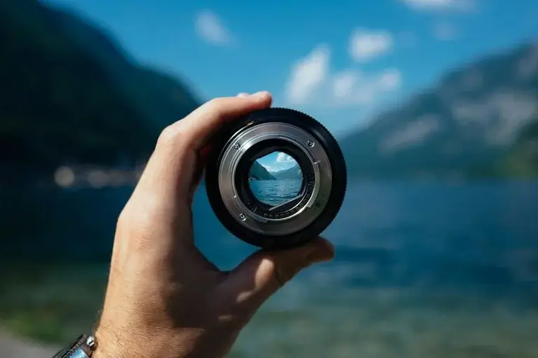 A hand holding a camera lens focusing on a natural mountain lake, representing the vision of making sustainability and responsible business a way of life.